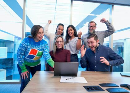 men and women sitting and standing by the table looking happy while staring at laptop